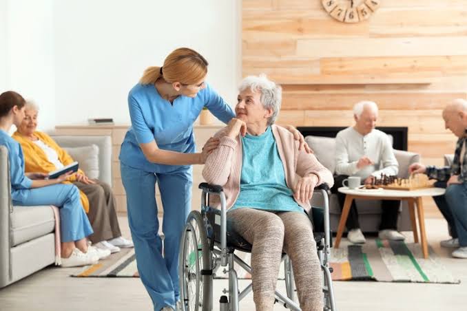 A caregiver leaning beside an older woman seated in a wheelchair in a shared sitting room