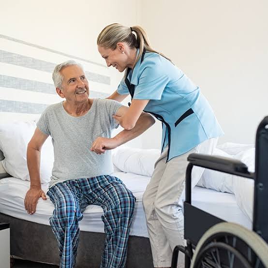 A caregiver helping an older man seated on the edge of his bed